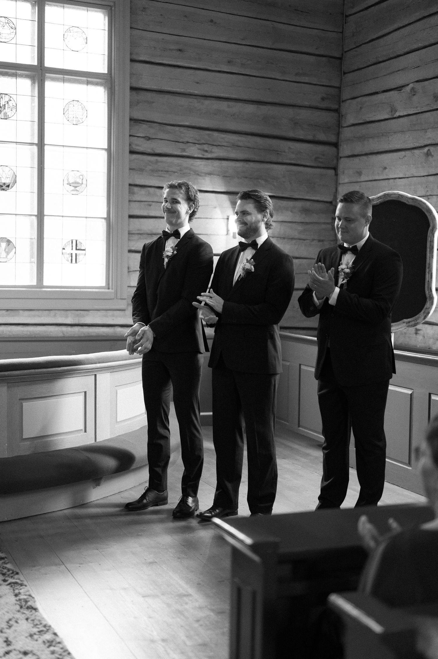 Groomsmen in black tuxedos pose in sequence along wooden wall with natural window light.