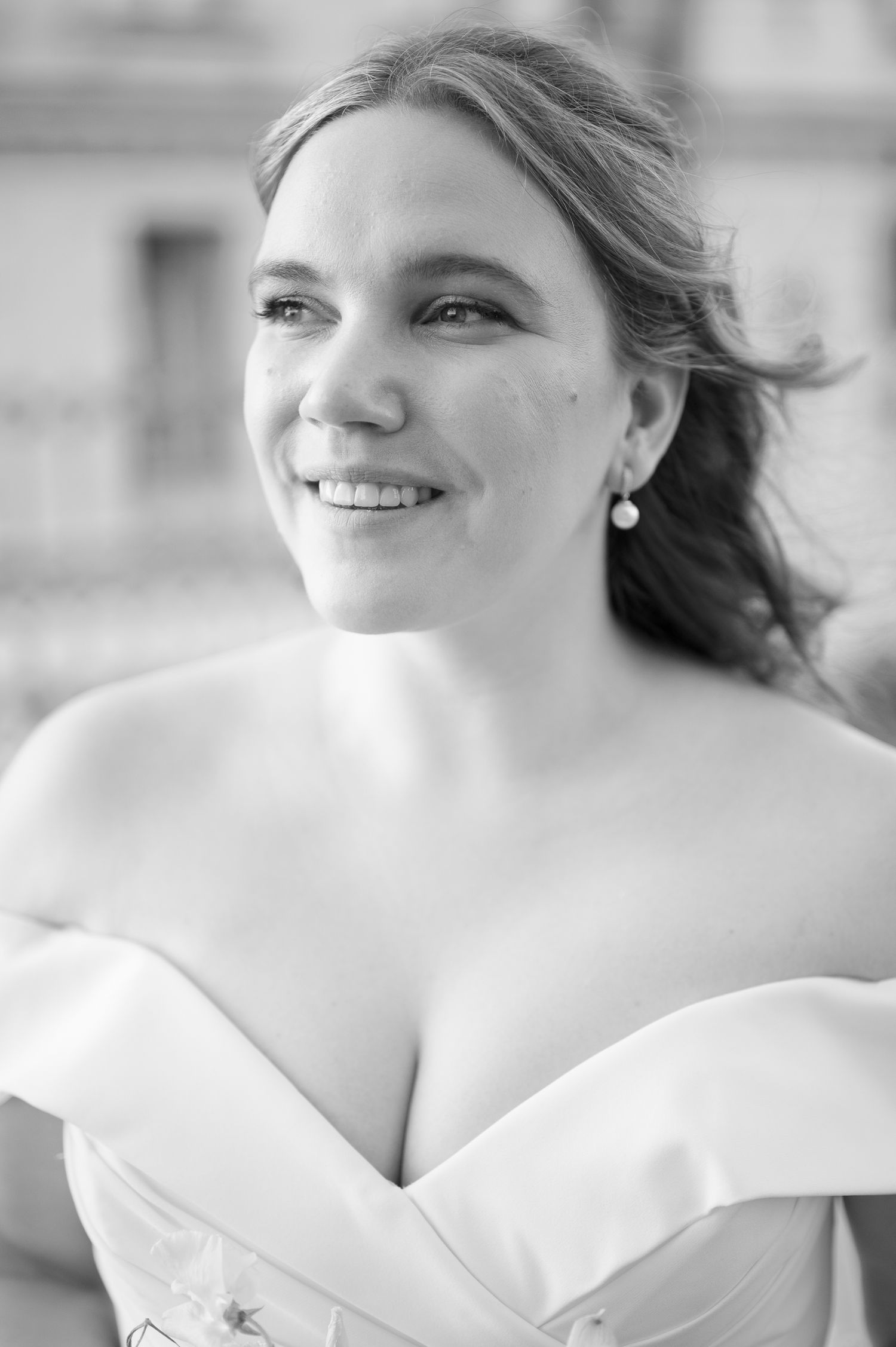 Black and white portrait of a smiling bride in an off-shoulder gown, wearing pearl earrings, with hair blowing in the wind.