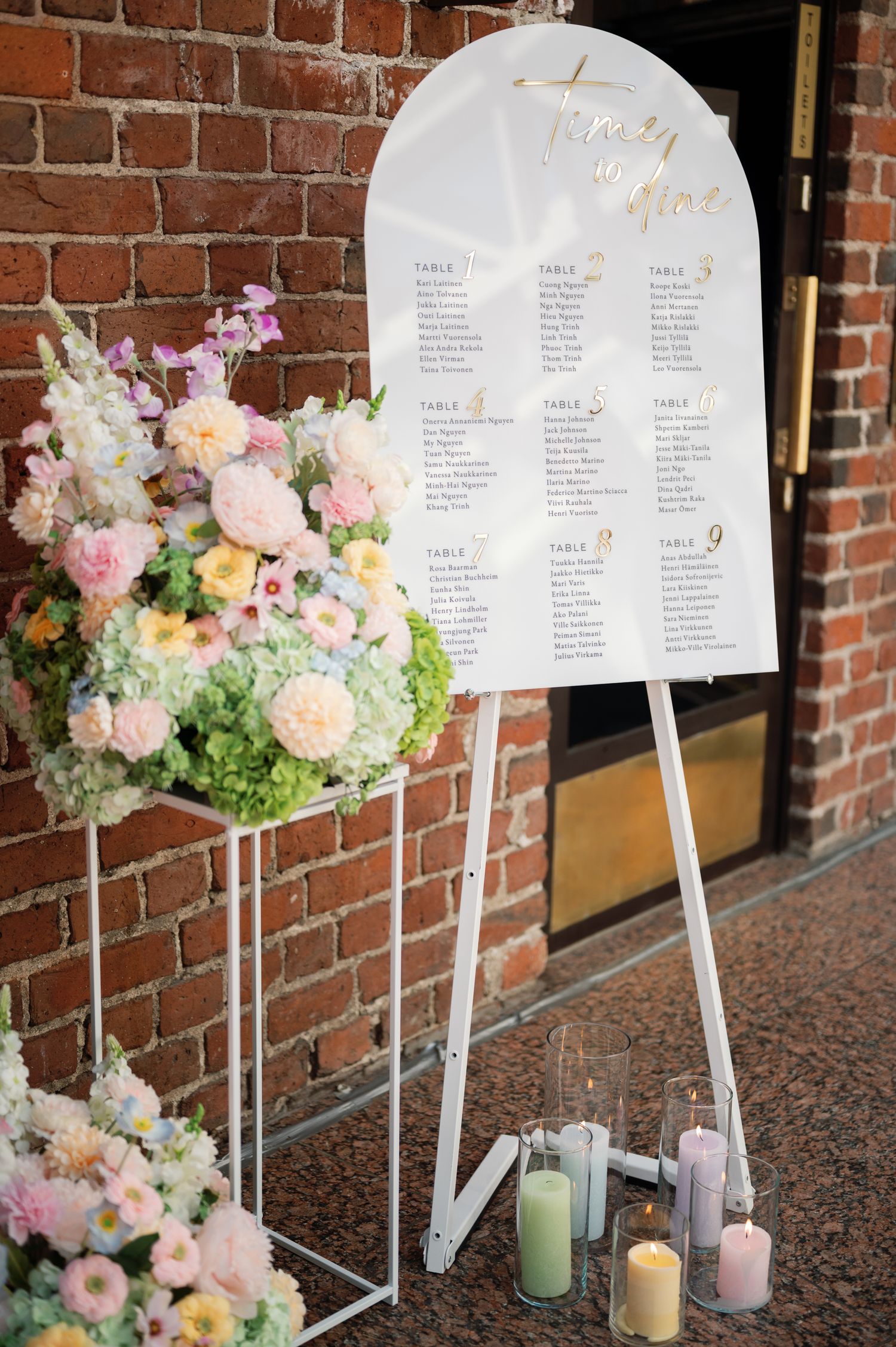 Close-up of wedding seating chart sign with gold calligraphy, pastel blooms, and colorful candles on white easel.