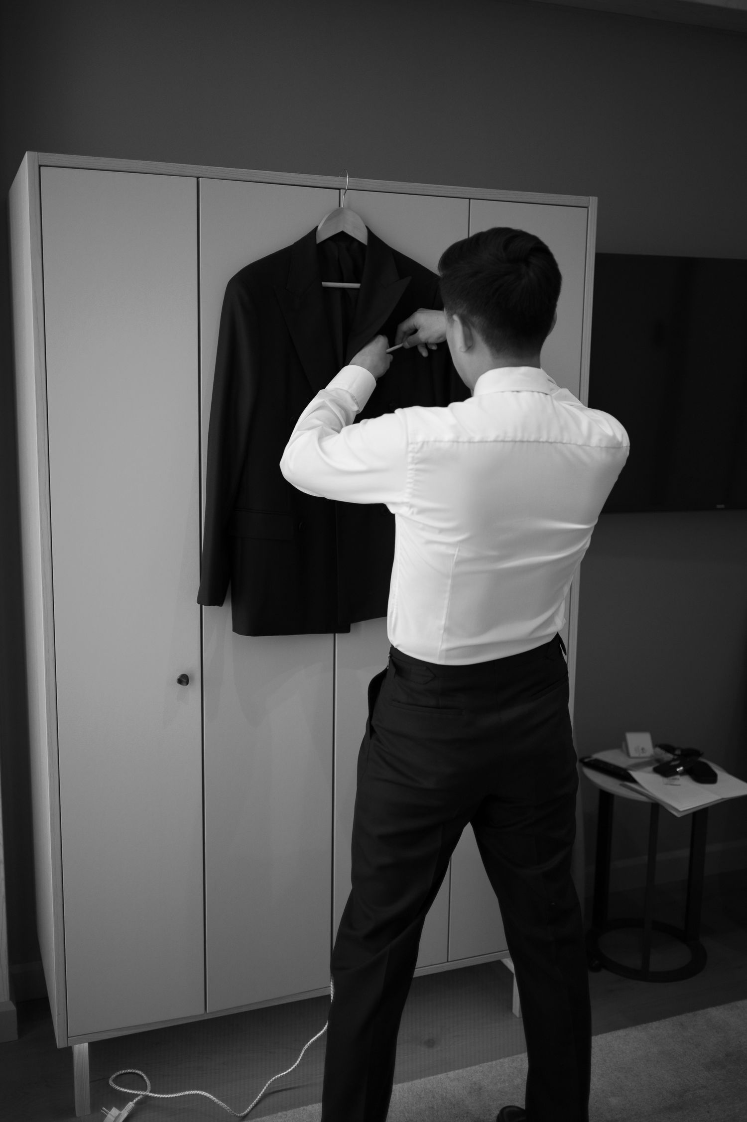 Man in white dress shirt preparing to put on a dark suit jacket hanging on a wardrobe in black and white.