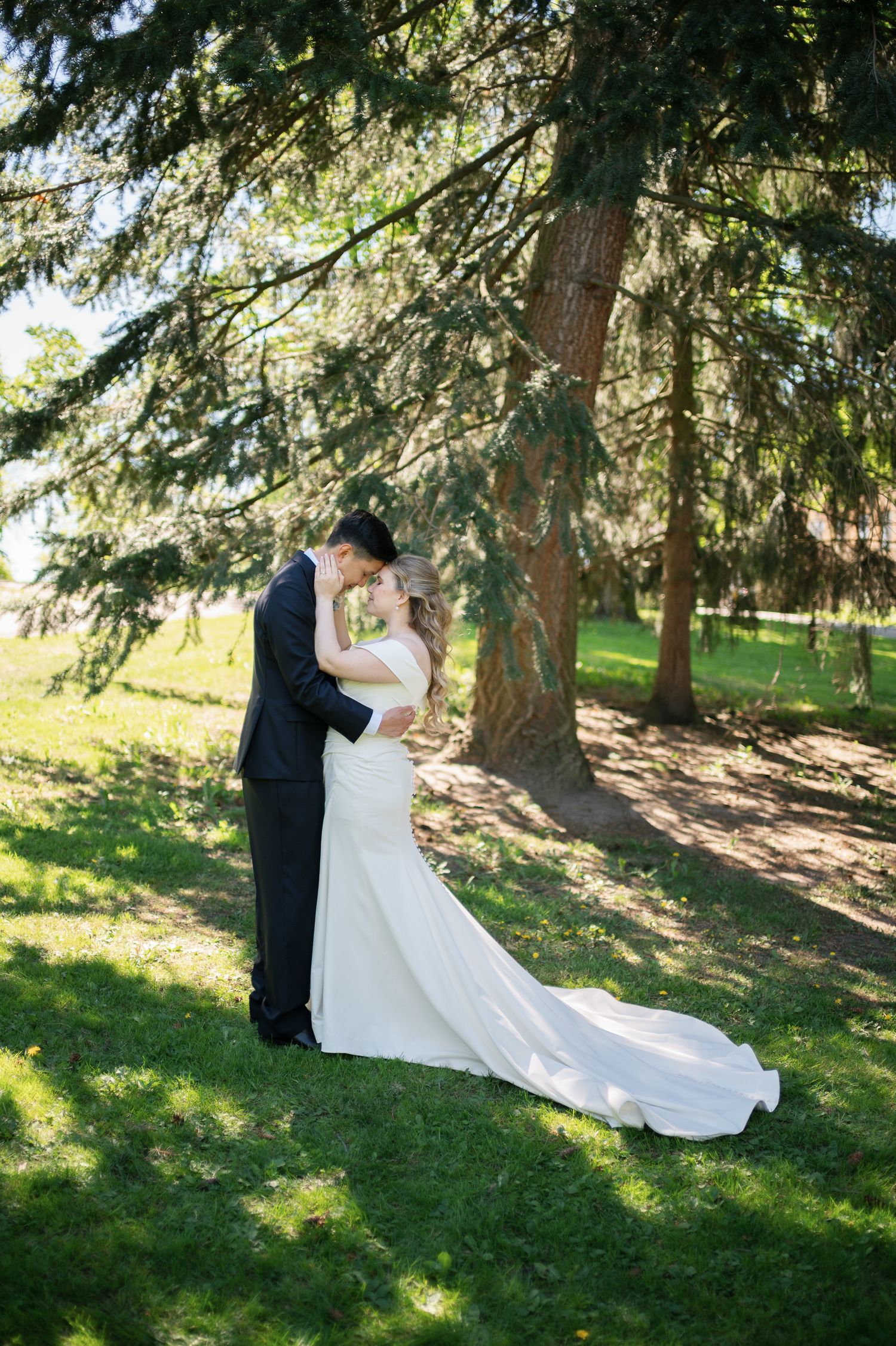 Bride in elegant white gown and groom in black tuxedo embrace under a large pine tree outdoors.