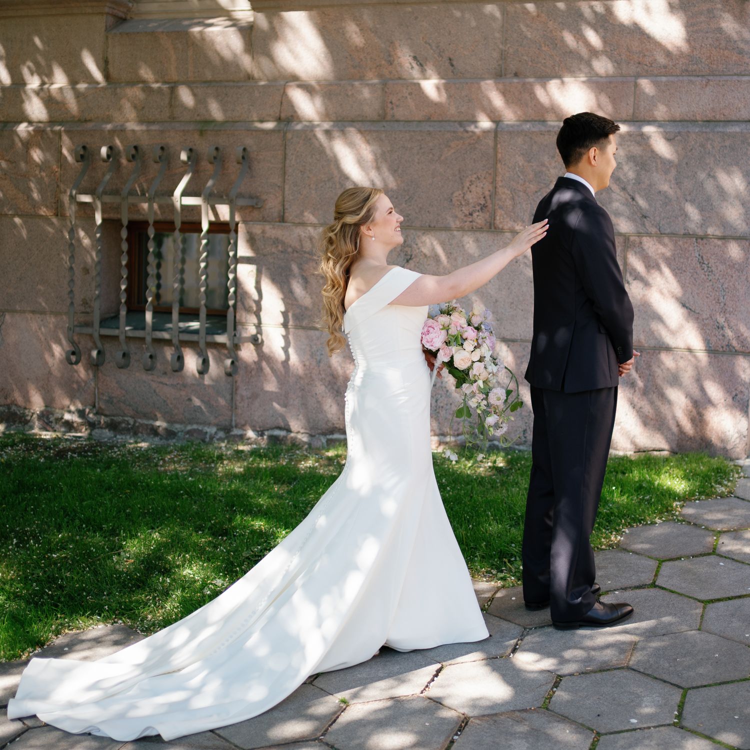 Bride in white gown touches groom's shoulder during first look at outdoor wedding venue with stone wall backdrop.
