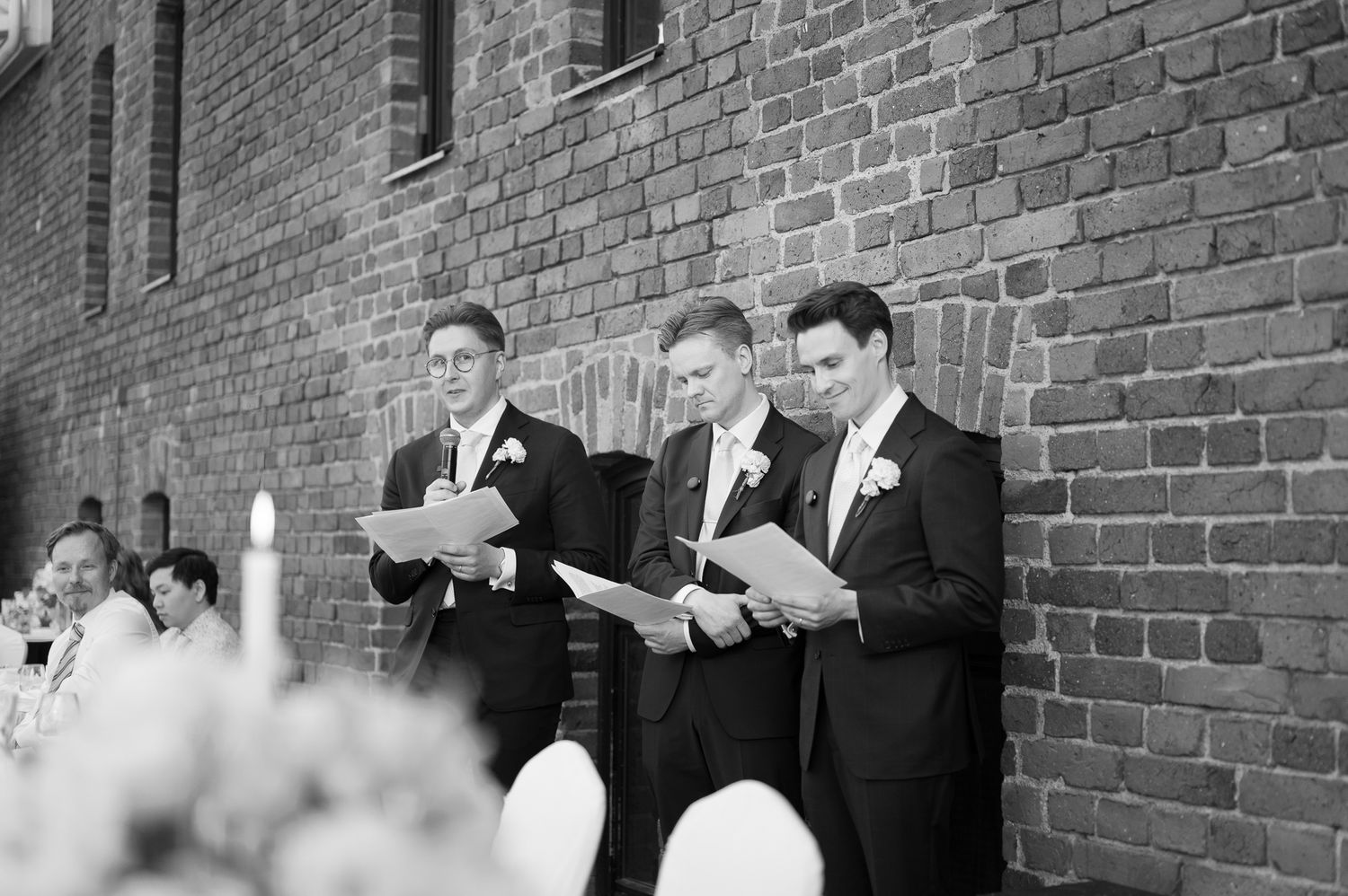 Three groomsmen in suits reading speeches at an outdoor wedding ceremony against a brick wall.