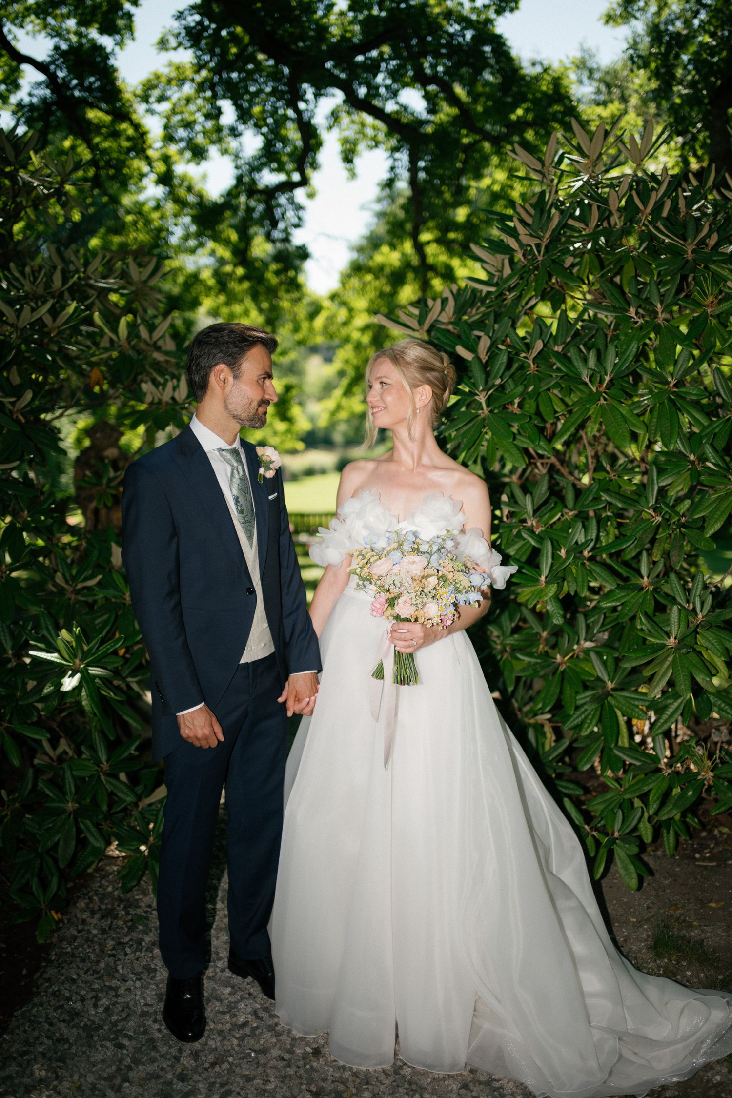 Bride and groom hold hands in lush garden, she wears off-shoulder gown holding pastel bouquet, he wears navy suit.