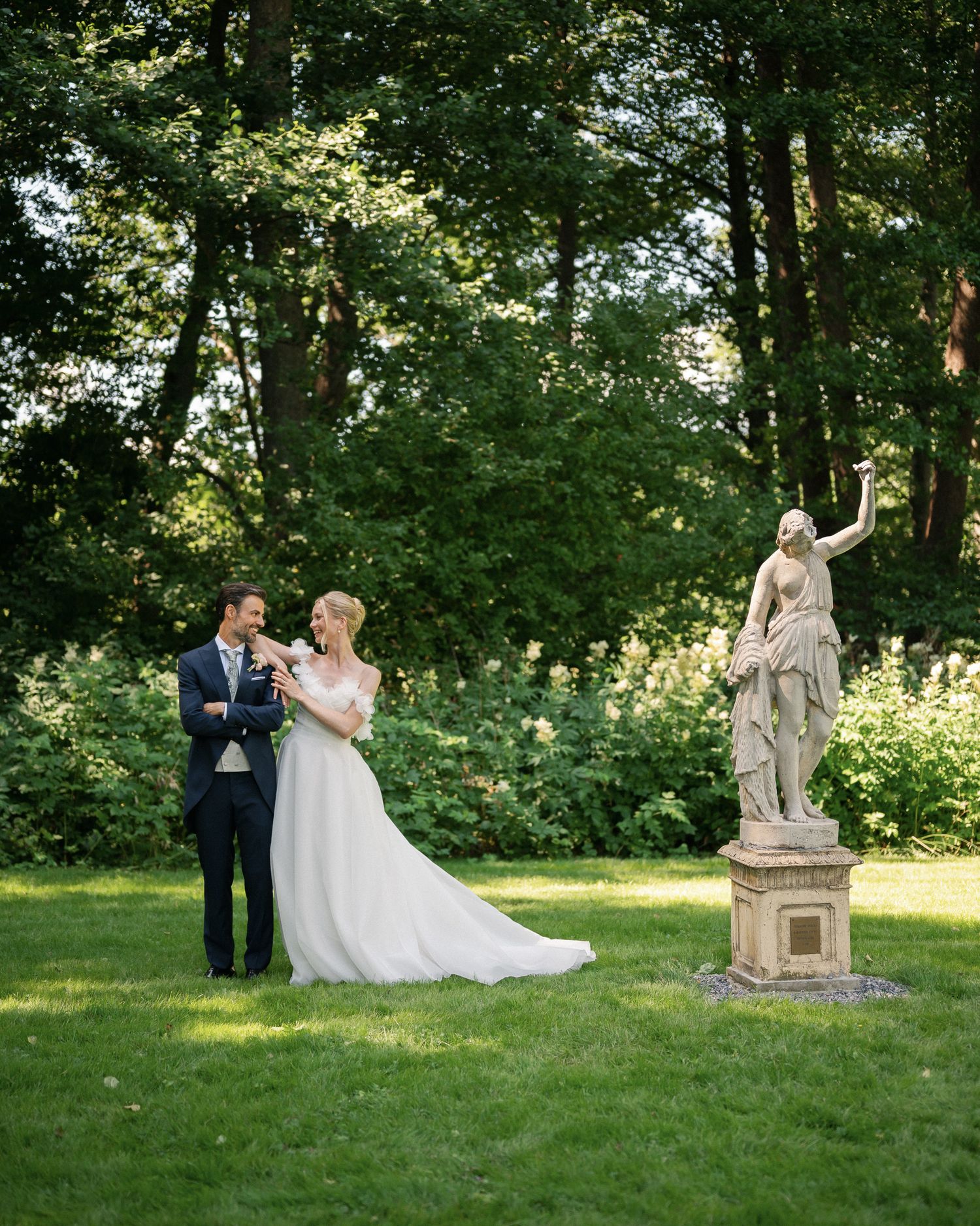 Bride and groom stand smiling in a lush garden beside an elegant stone statue on their wedding day.