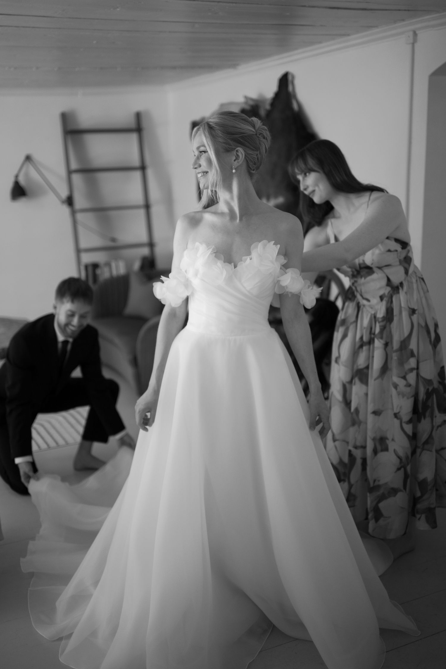 Black and white photo of a bride in a floral off-shoulder gown being helped by a bridesmaid.