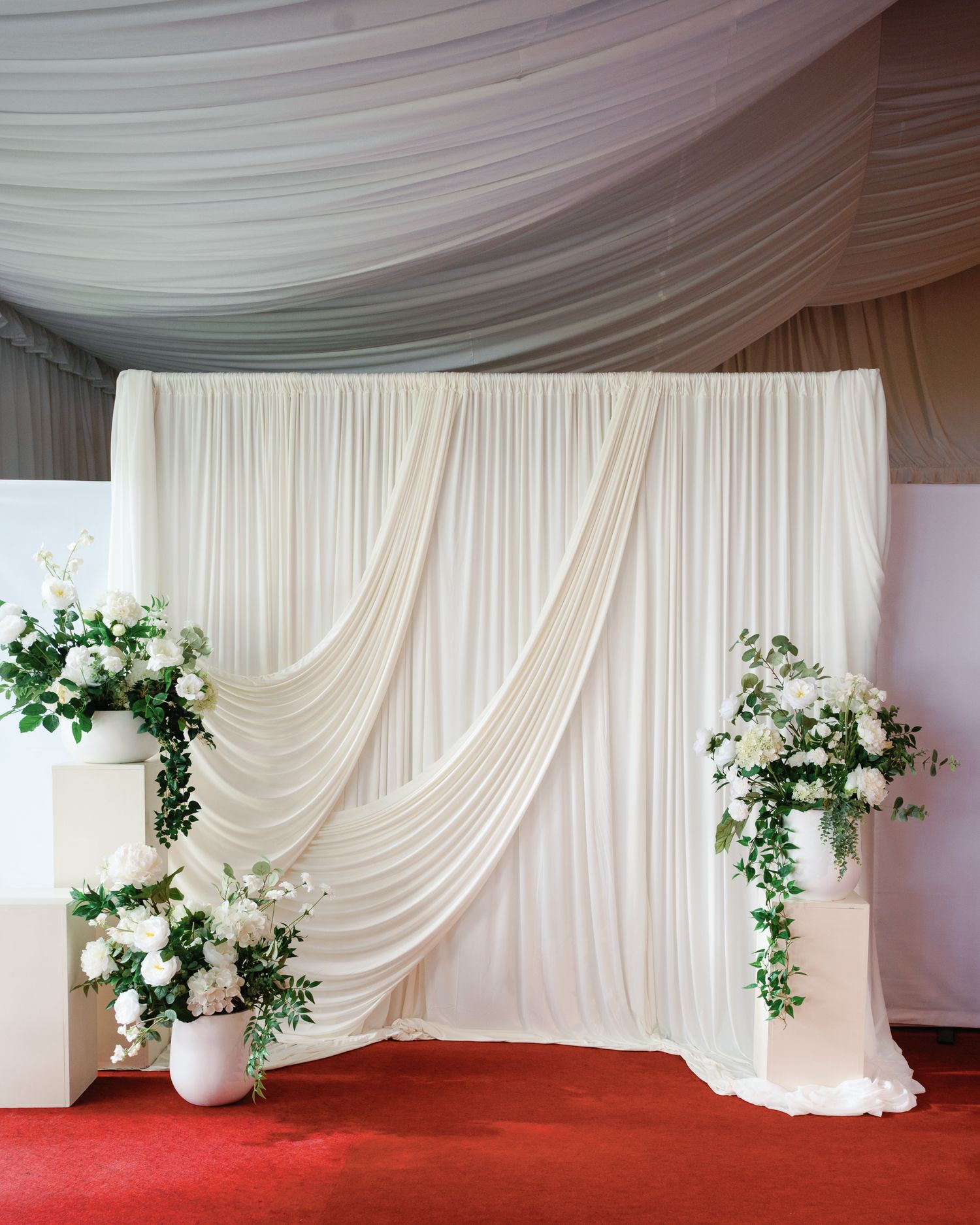 Elegant white draped fabric backdrop with white floral arrangements on pedestals, perfect for weddings.