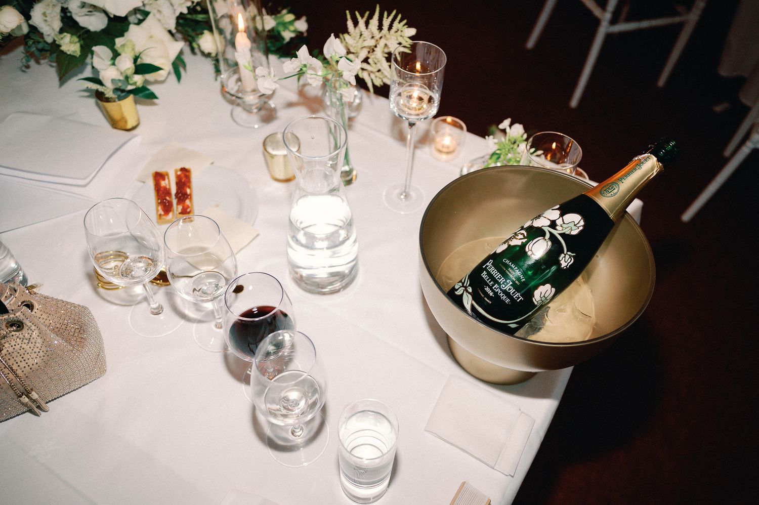 Elegant wedding reception table with champagne in gold ice bucket, candles, and glassware on white linen.