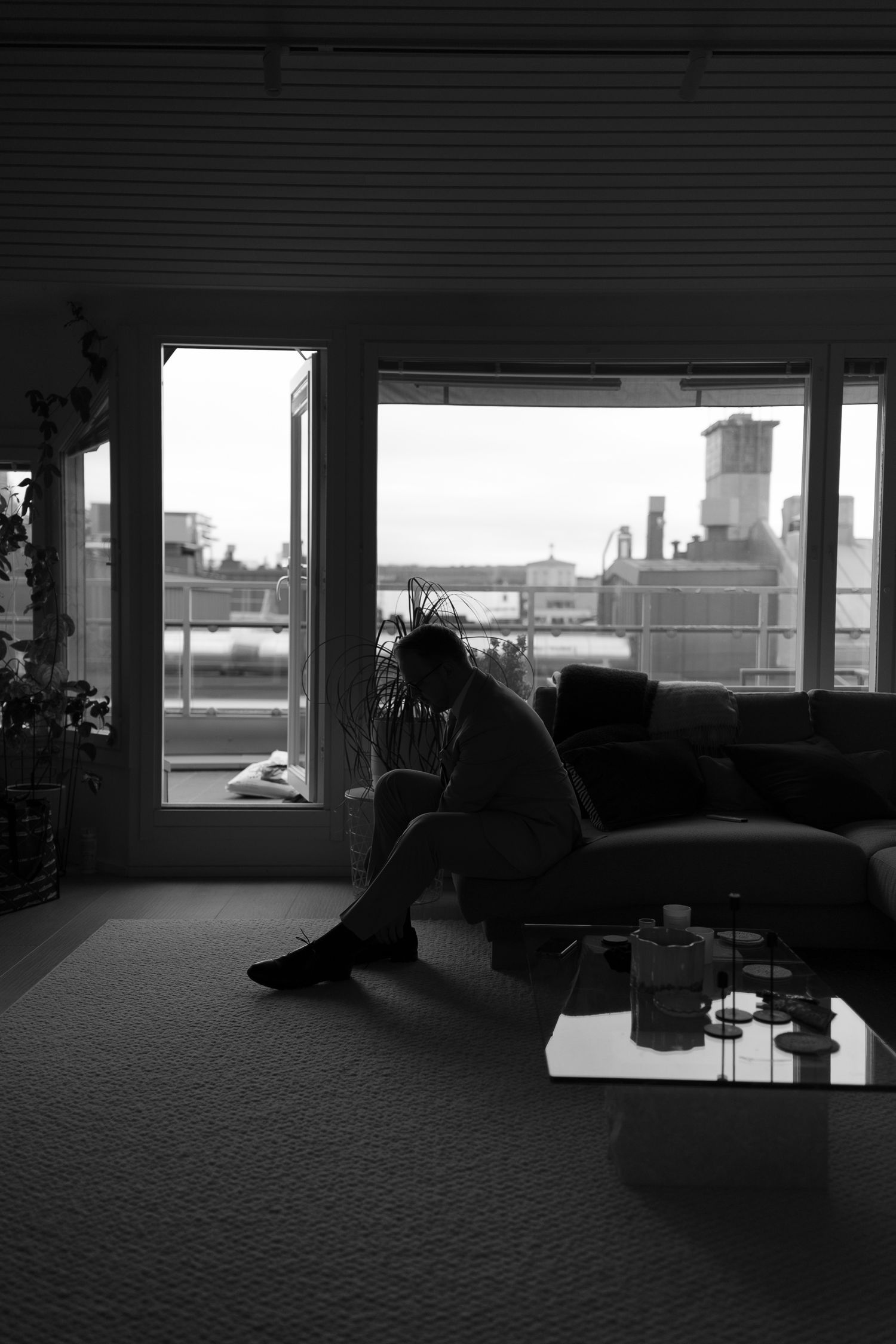 Black and white photo of a person sitting alone on a couch, head bowed, in a dimly lit room with large windows.