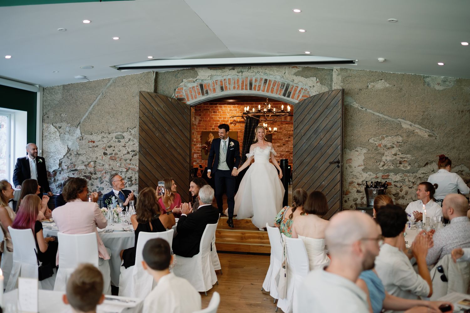 Bride and groom make grand entrance into elegant rustic wedding reception hall with stone walls and seated guests.