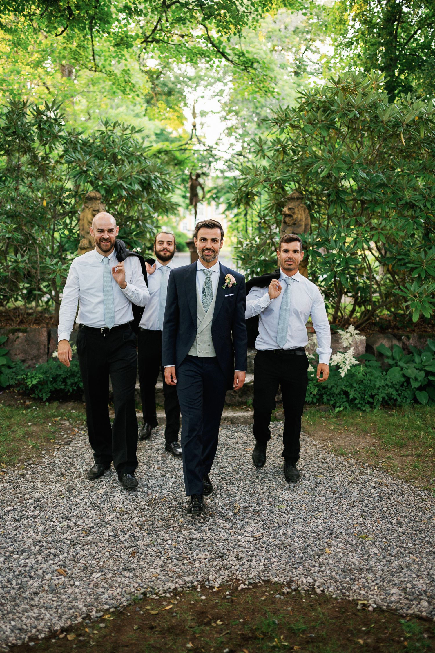Groom in navy suit leads groomsmen in white shirts carrying jackets on gravel path surrounded by lush greenery.