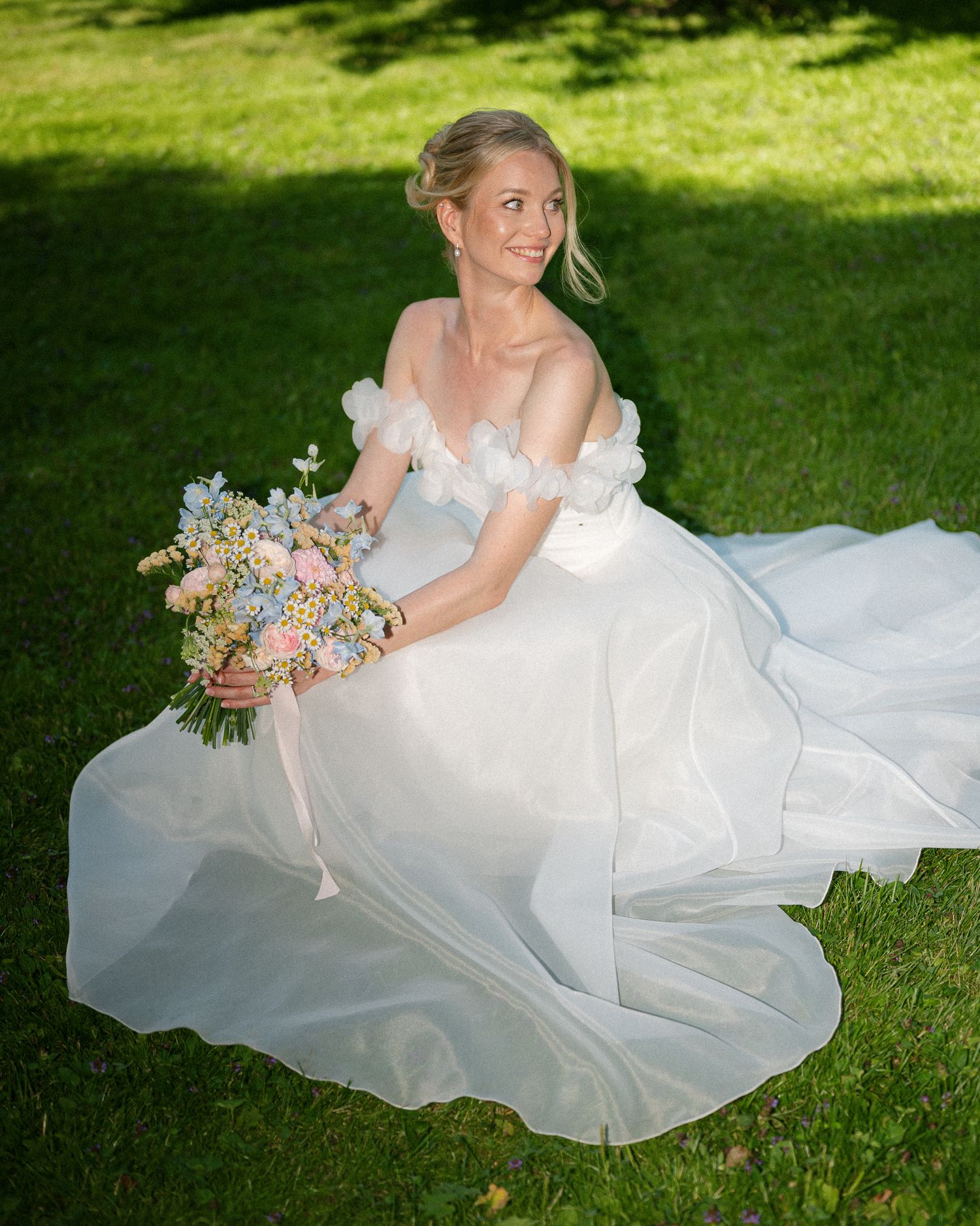 Bride in off-shoulder white gown sits on green grass holding pastel blue and pink bouquet, smiling outdoors.