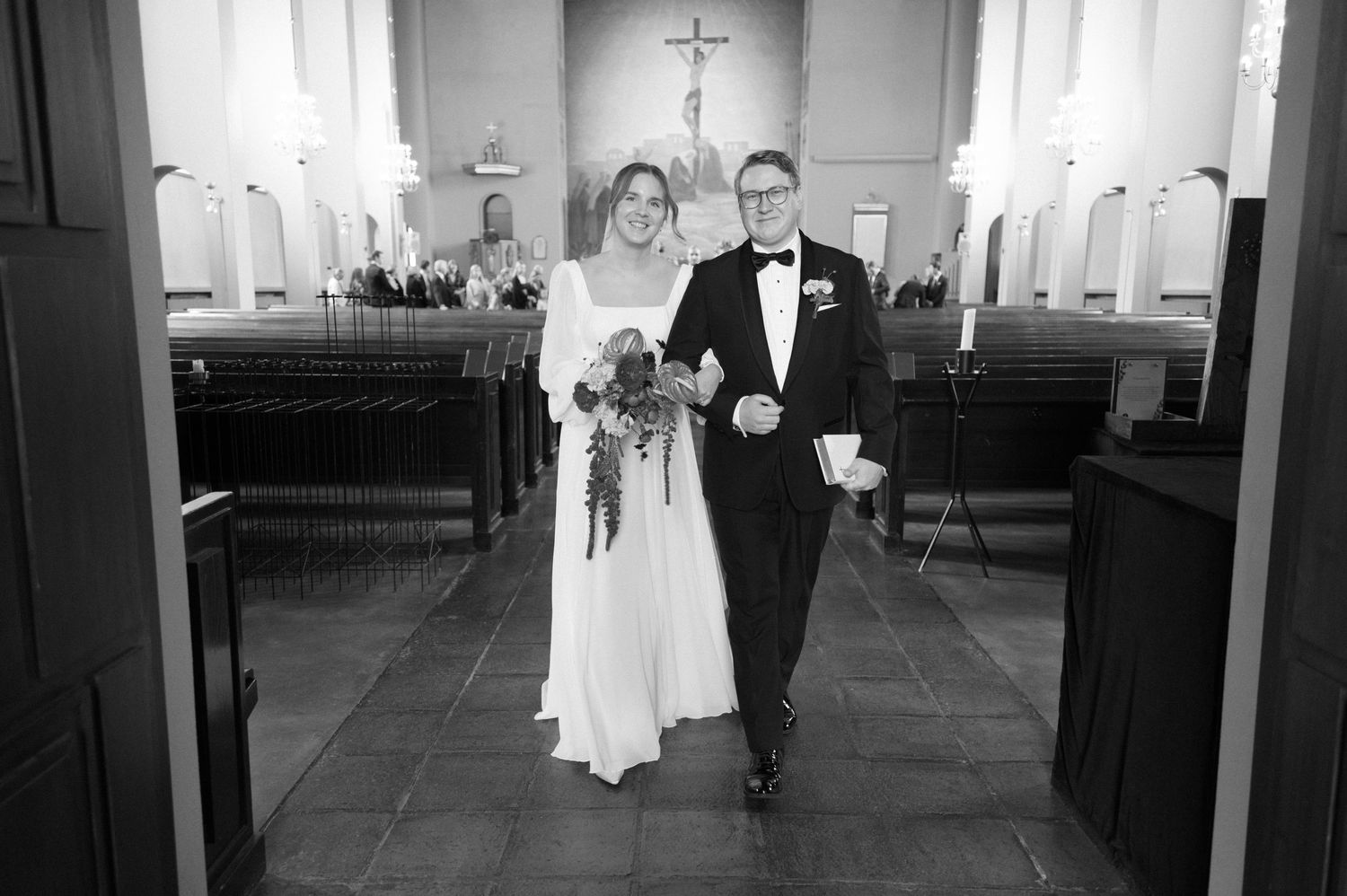 A wedding couple walks down the aisle of a church sanctuary with white walls and wooden pews.
