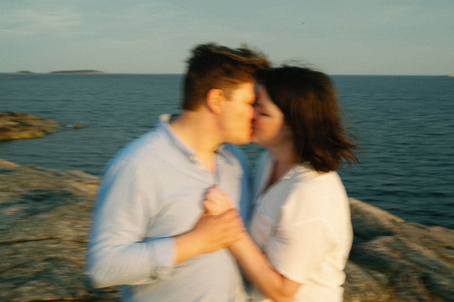 Romantic couple in white clothing embracing on rocky coastal cliffs at sunset.