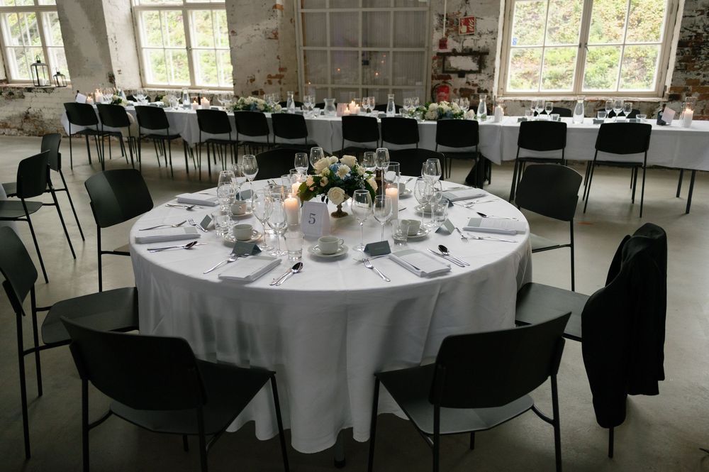 Round dining tables with white tablecloths set for a wedding reception in an industrial venue.