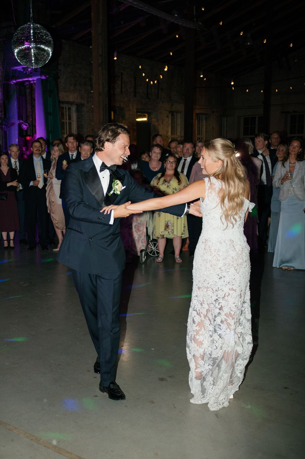 A couple performs an elegant dance dip move on a dimly lit dance floor surrounded by wedding guests.