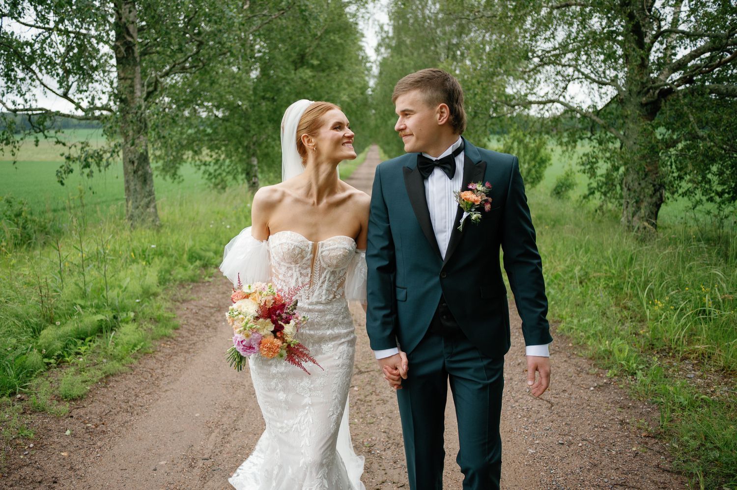 A romantic wedding couple strolls along a tree-lined country path in elegant attire.