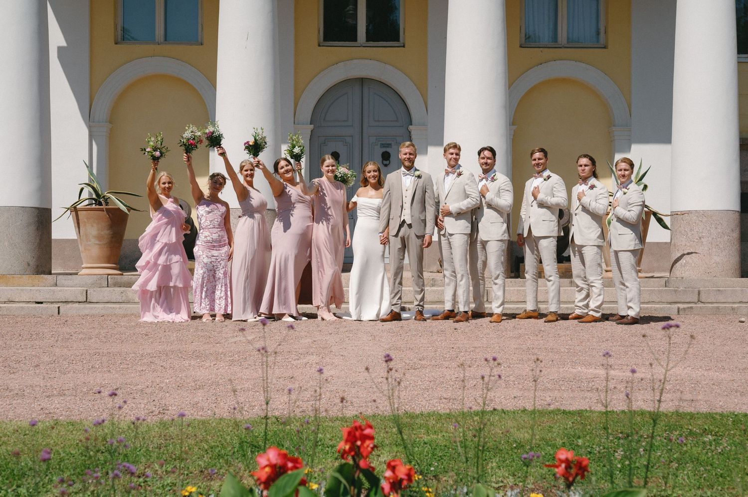 Wedding group poses playfully on stone steps of classical building with white columns.