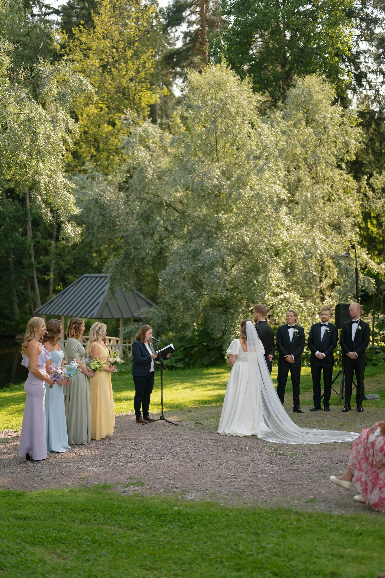 Wedding party assembles for outdoor ceremony against backdrop of lush green trees.