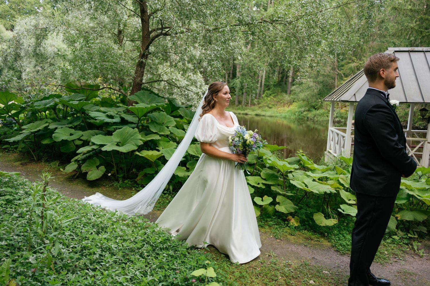 A couple shares a moment on a garden path with flowing white dress and elegant attire.