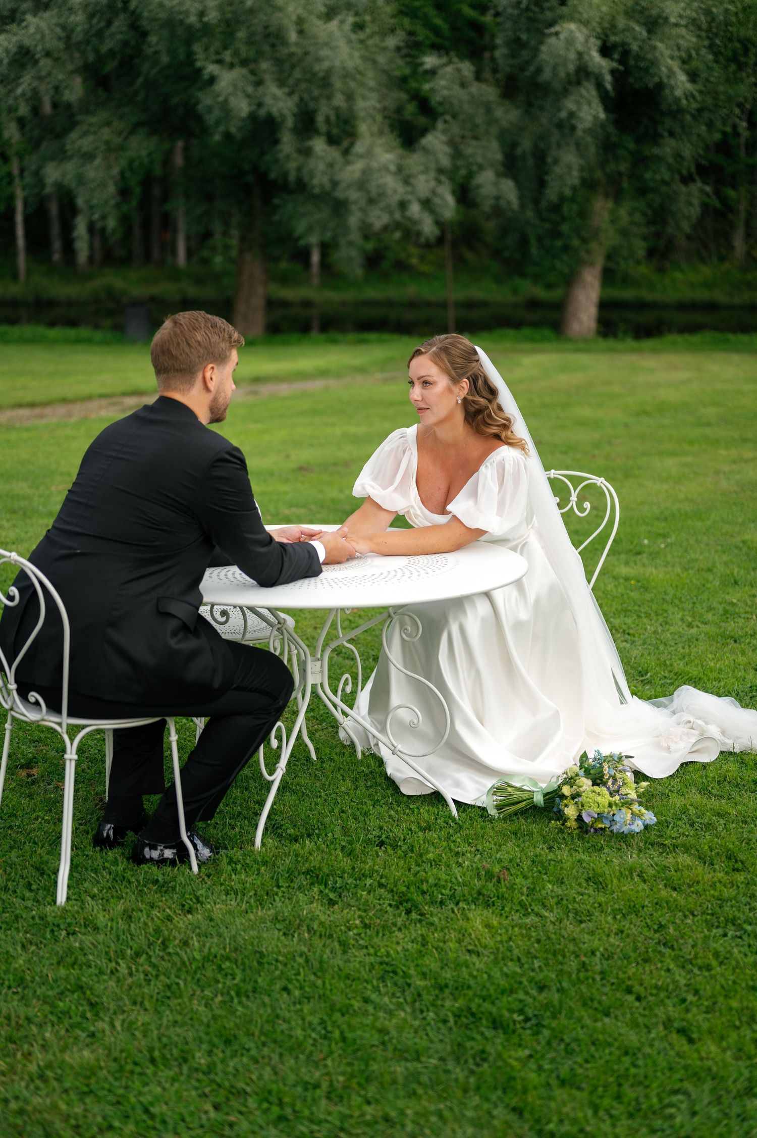 Wedding couple reclining on white garden chairs in a scenic park setting.