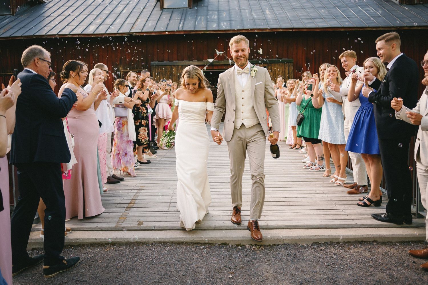Wedding party walks down wooden deck aisle while guests throw confetti in celebration.
