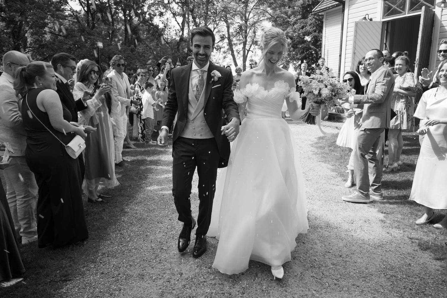 Black and white photo of a bride and groom walking hand in hand through cheering wedding guests outdoors.
