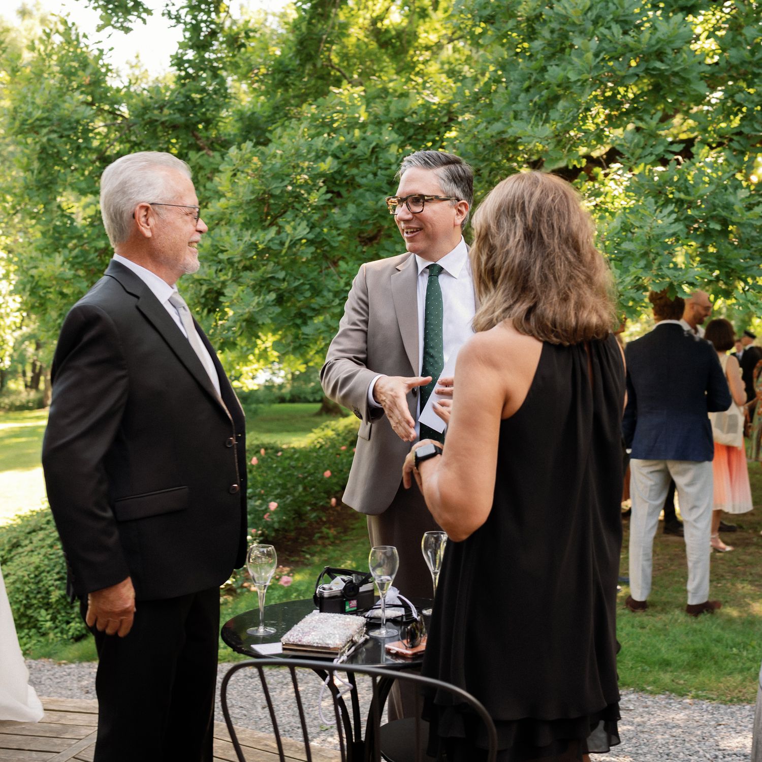 Three elegantly dressed guests converse outdoors at a garden reception, surrounded by lush green trees.