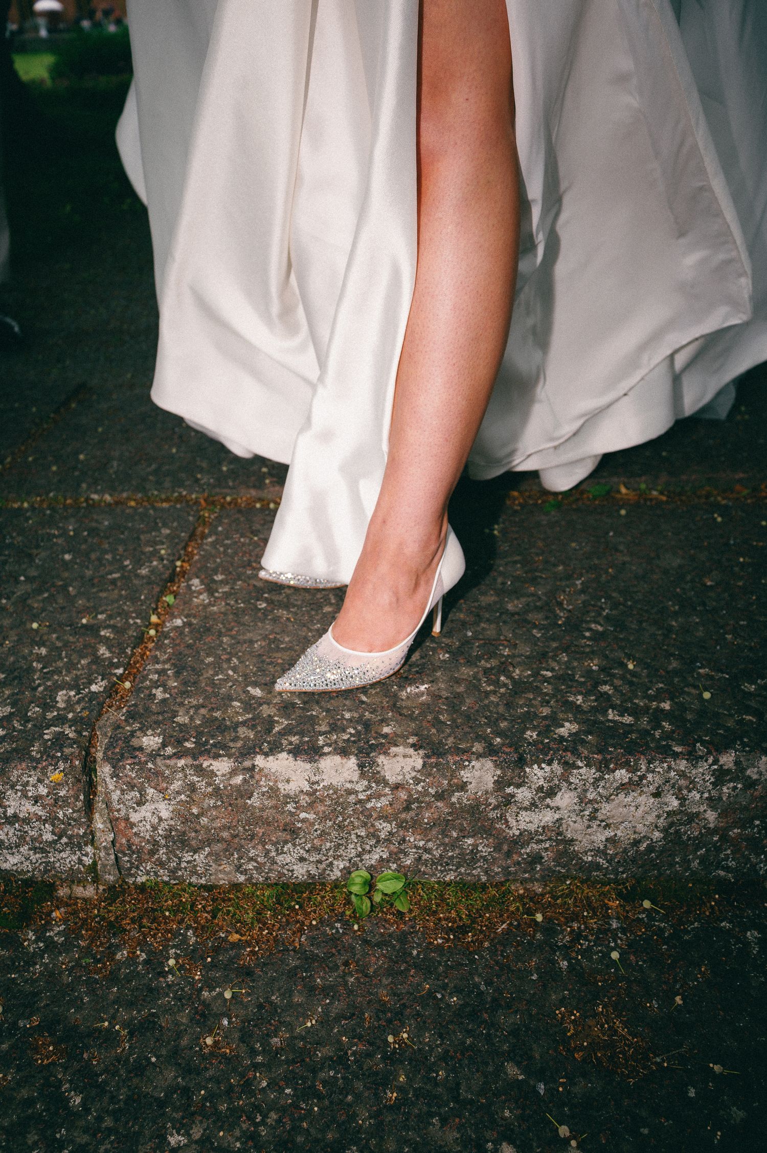 Bride in white gown stepping off curb wearing sparkling silver glitter pointed-toe heels on wet pavement.