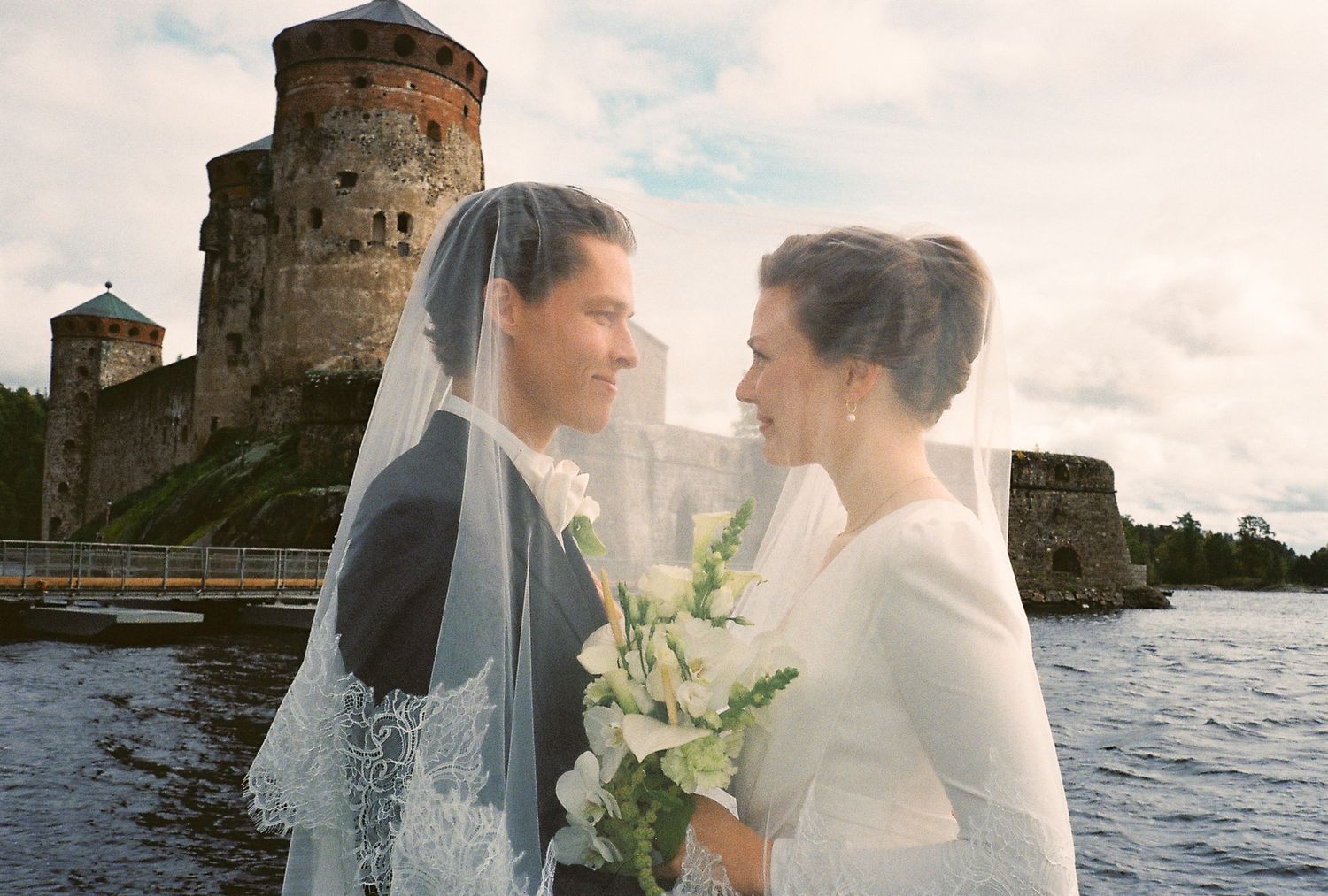 Bride and groom share a romantic moment by a lake with a medieval castle backdrop on their wedding day.