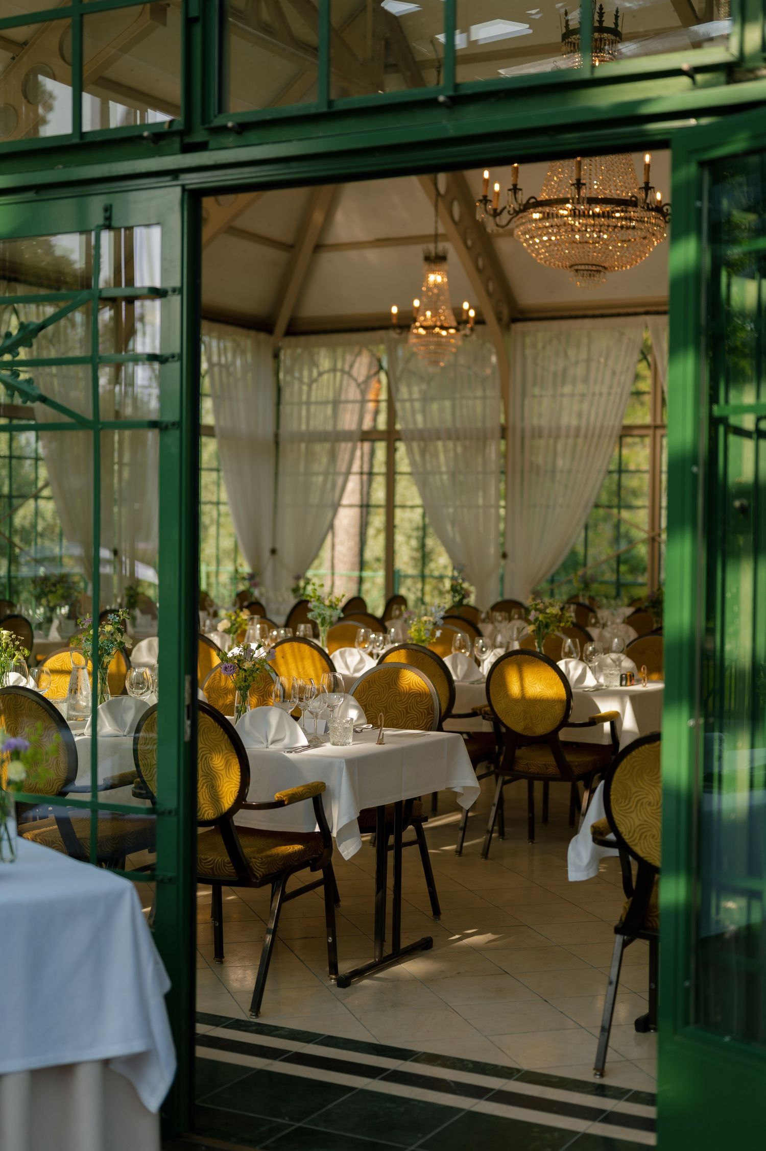 Elegant dining room with crystal chandeliers viewed through green-framed glass doors.