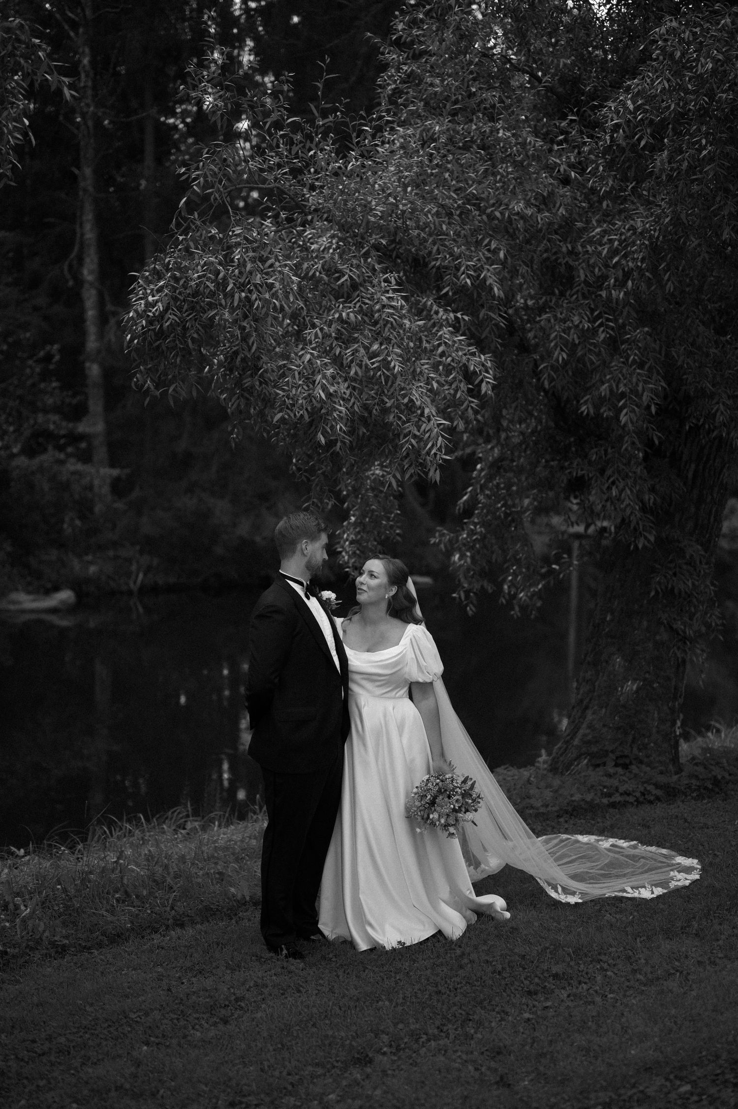 Black and white wedding portrait with couple standing near trees and water.