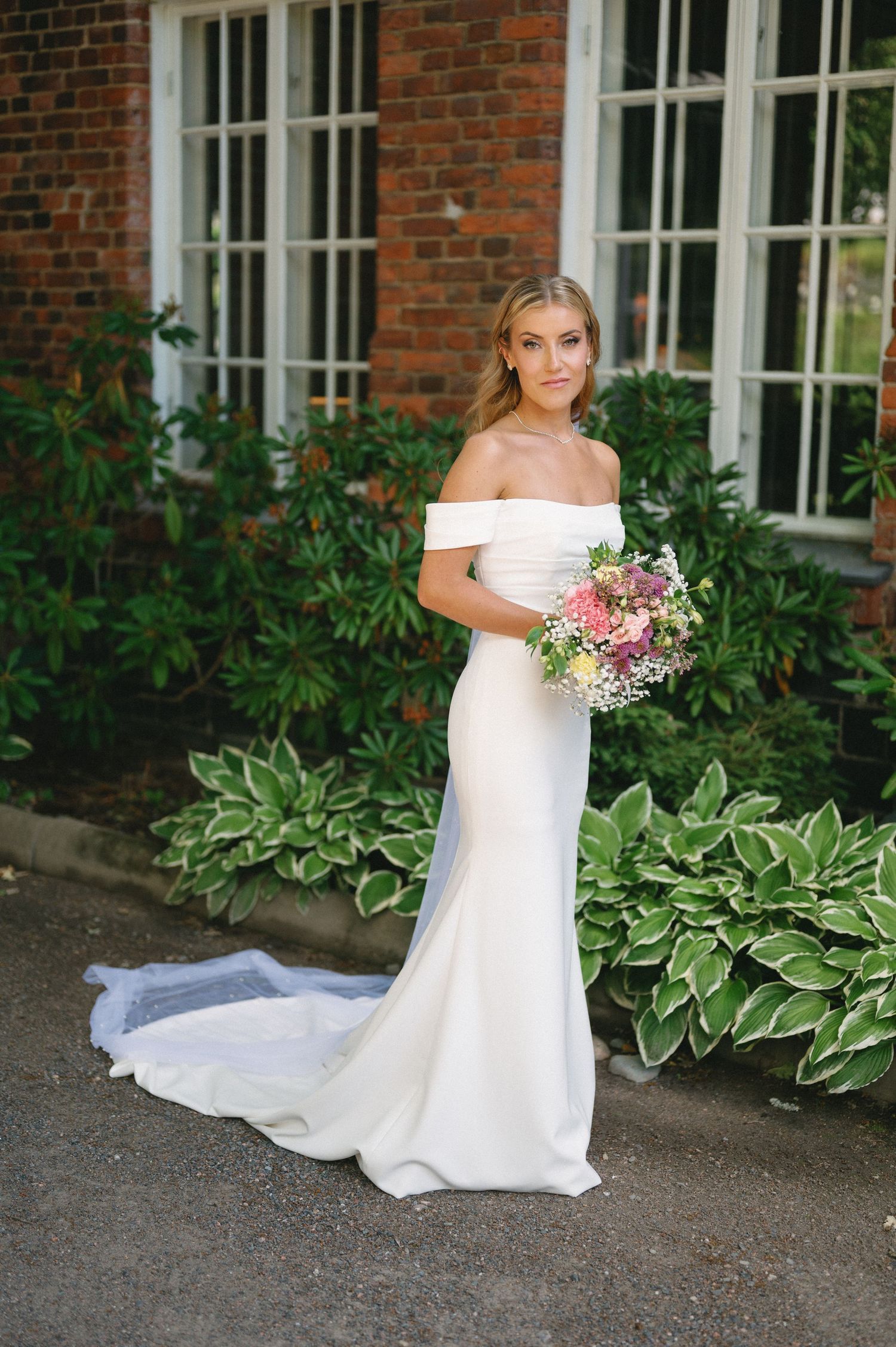 Wedding dress detail with flowing train against red brick building and garden backdrop.