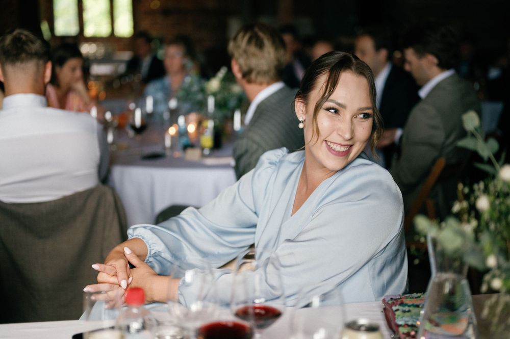 Guest in light blue jacket smiles at wedding reception table with wine glasses.
