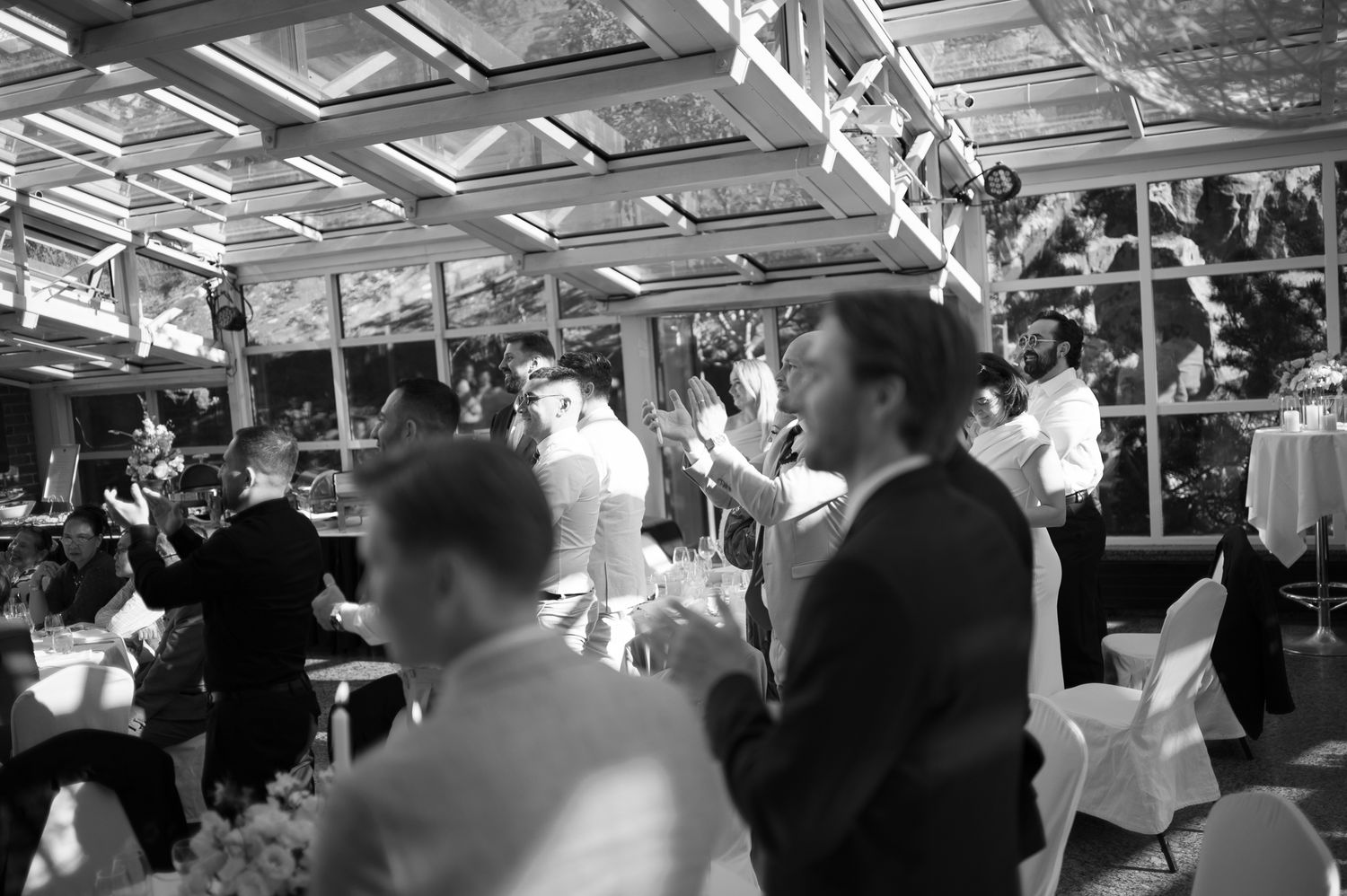 Black and white photo of elegantly dressed guests mingling at a wedding reception in a glass-roofed venue.