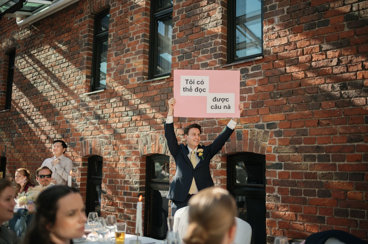 Man in suit holds pink sign above head at outdoor wedding reception near brick building.