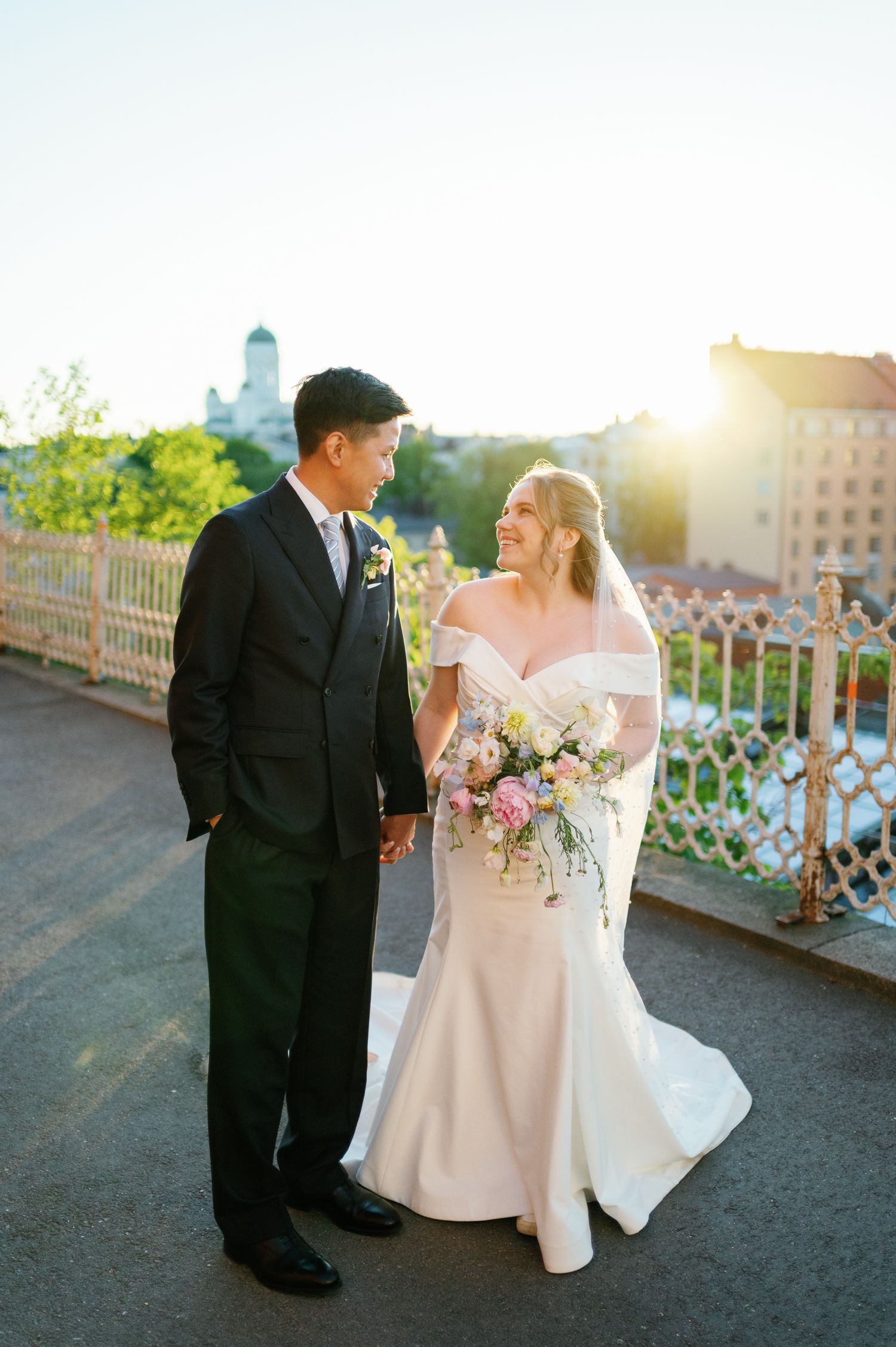 Bride and groom hold hands on a sunlit bridge, she in a white off-shoulder gown with bouquet, he in a black tuxedo.