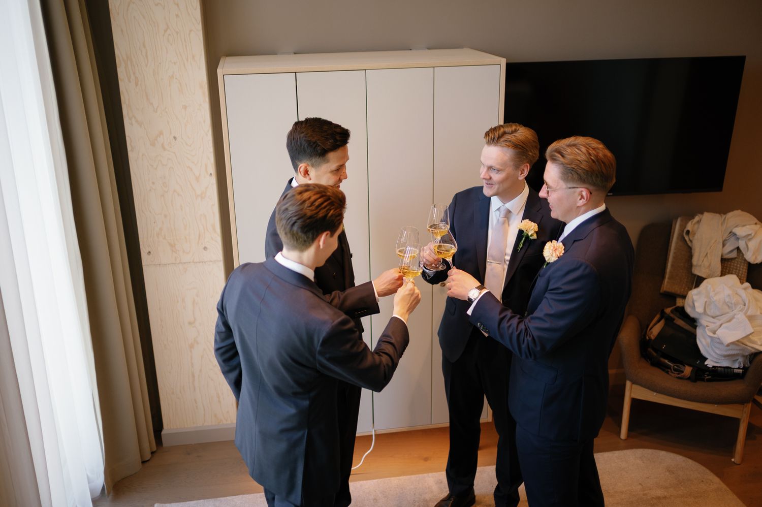 Three groomsmen in navy suits toasting with champagne glasses in a hotel room before a wedding ceremony.