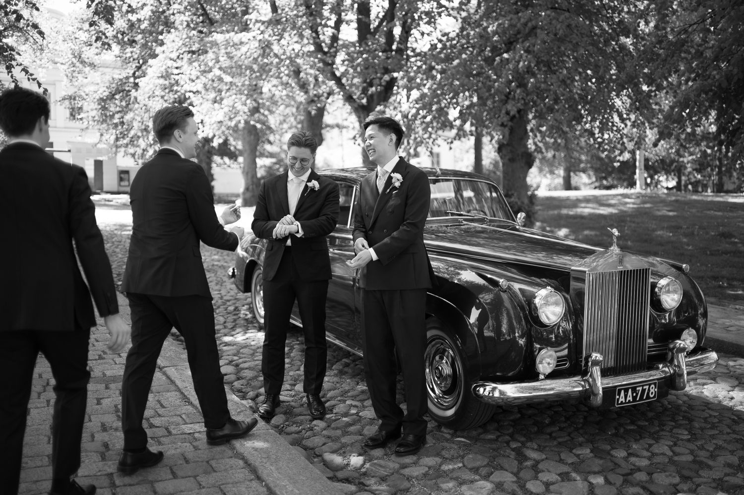 Men in suits greet each other beside a classic vintage Rolls-Royce car in a black and white wedding photo.