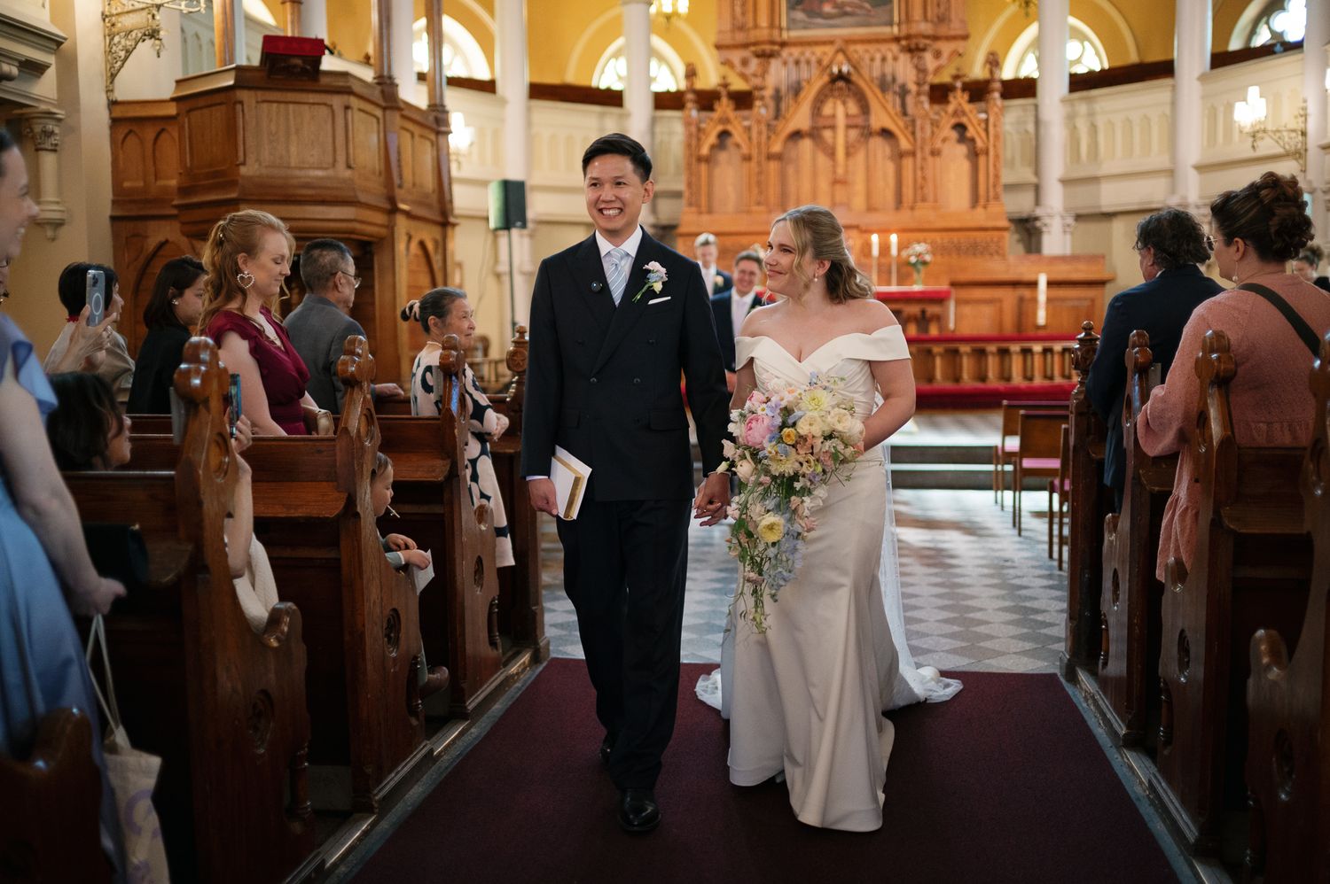 Newlywed couple walking down church aisle after wedding ceremony, bride in white gown holding bouquet, groom in dark suit.