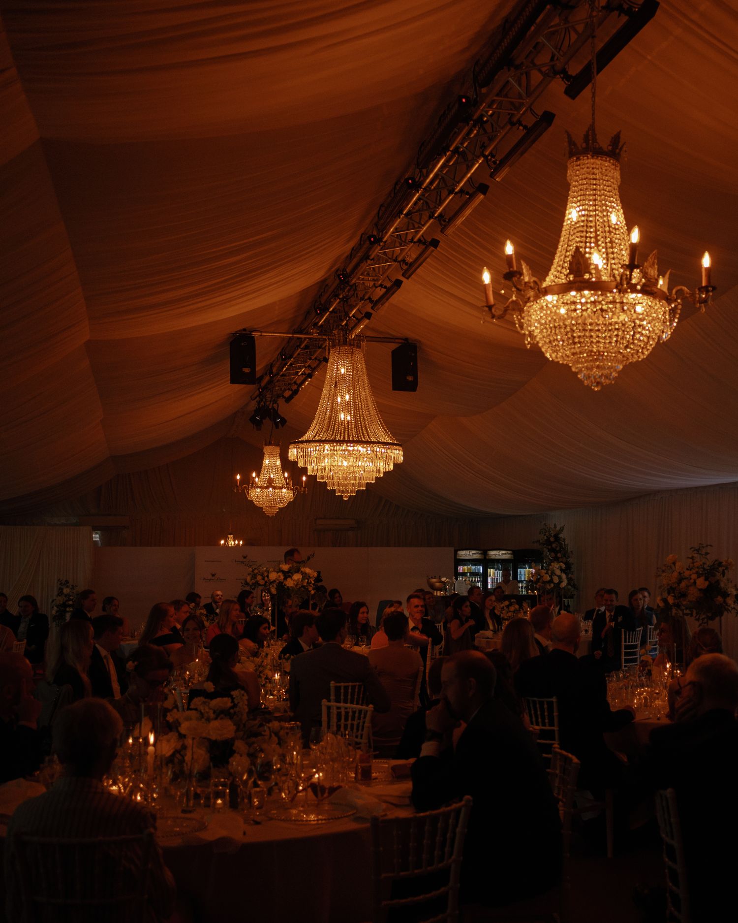 Elegant wedding reception in a draped tent with crystal chandeliers, candlelit tables, and guests gathered beneath warm golden lighting.