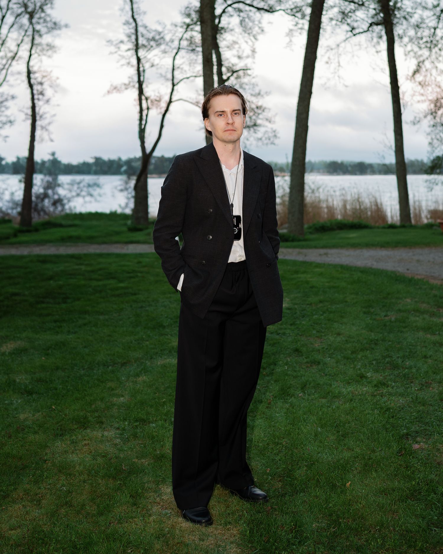 Young man in black suit standing on green lawn near lake with trees in background.