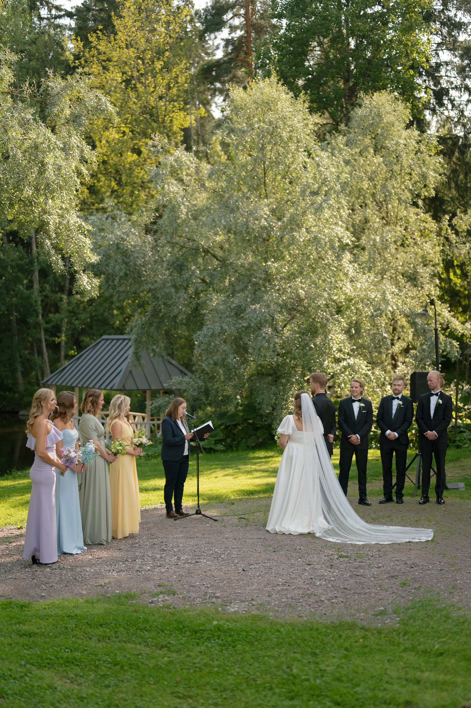 Wedding party stands in formation on grassy outdoor area surrounded by lush green trees during summer ceremony.