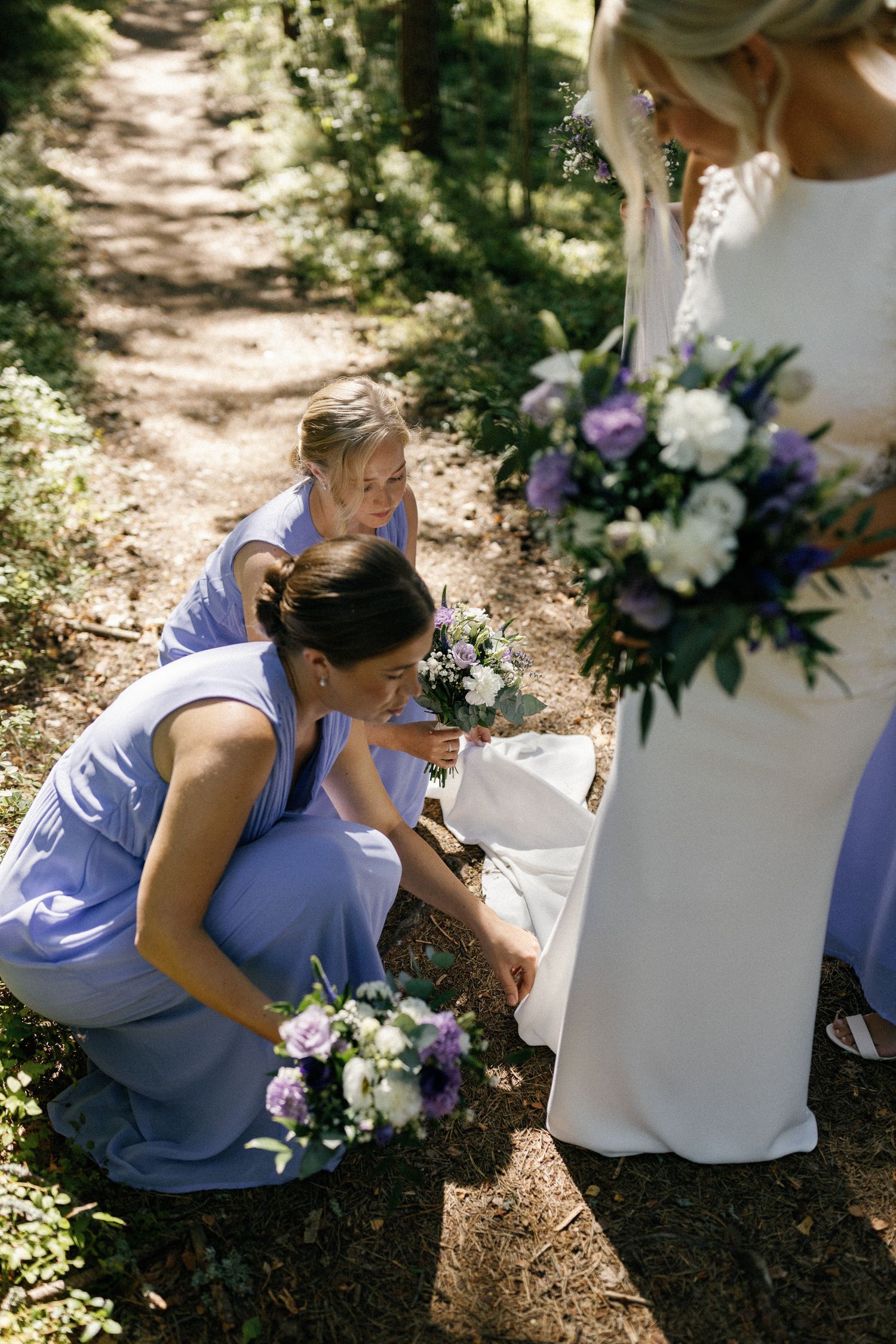 Bridesmaids in light blue dresses adjust wedding attire in a forest setting.