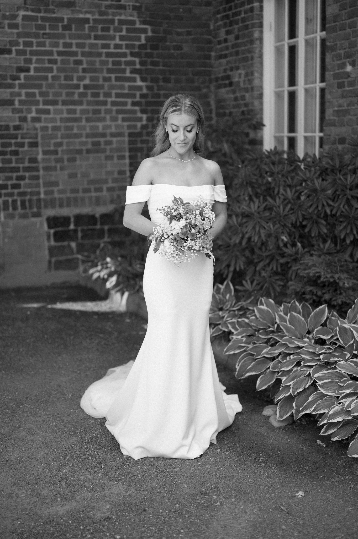 Black and white portrait showing off-shoulder wedding dress details near garden setting.