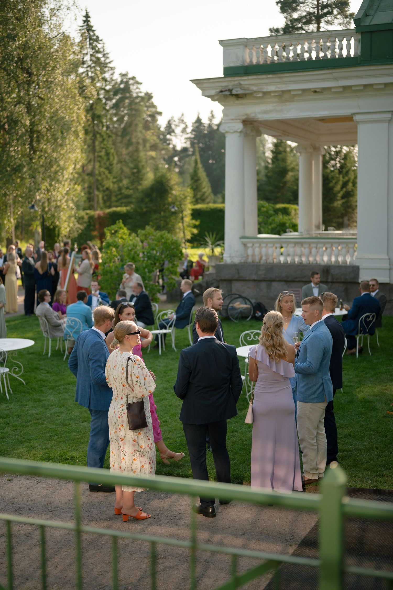 Wedding guests mingling on a lawn near a classical white columned pavilion at sunset.