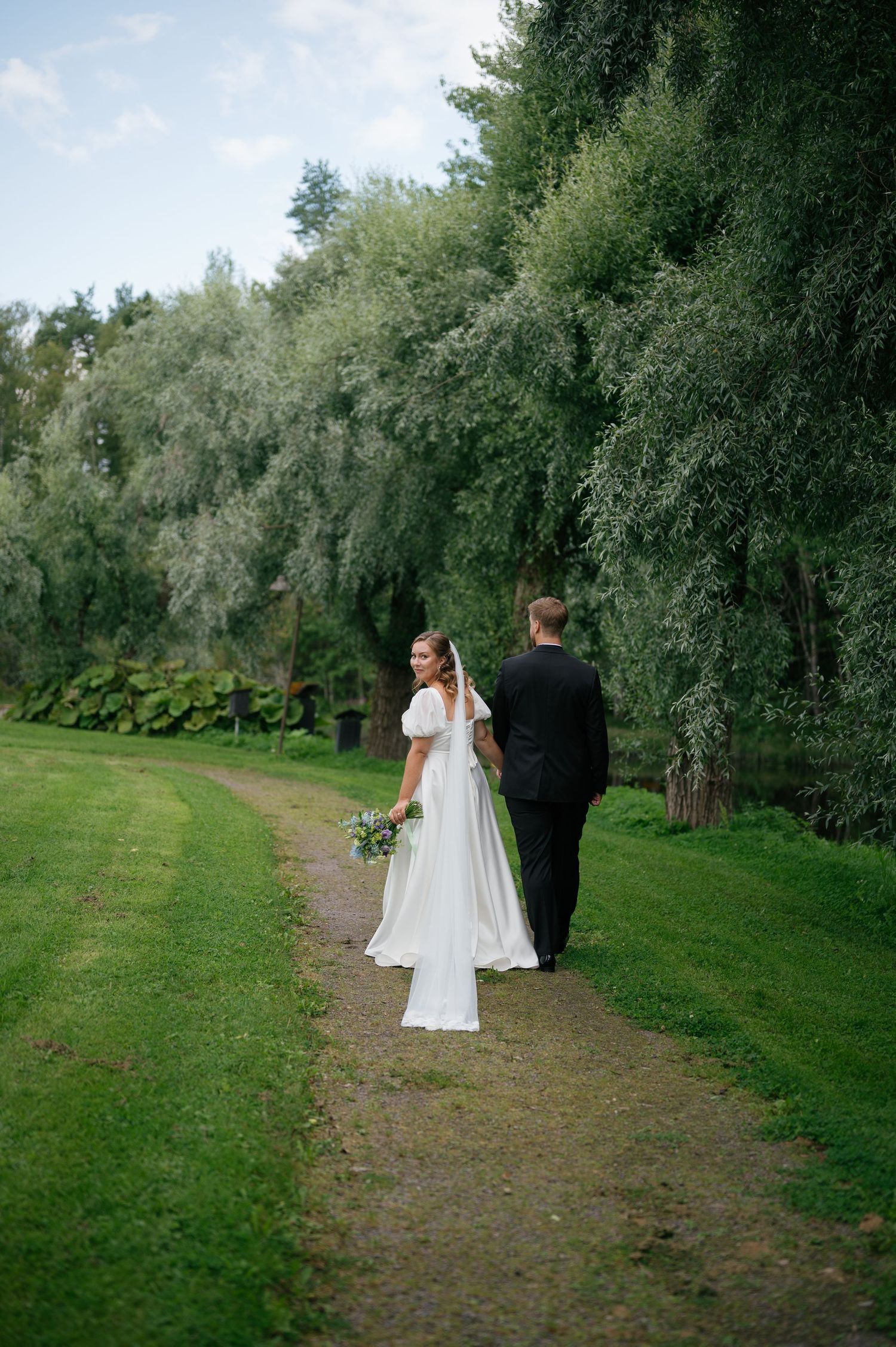 Couple walking along a tree-lined dirt path in a garden setting during a wedding photoshoot.