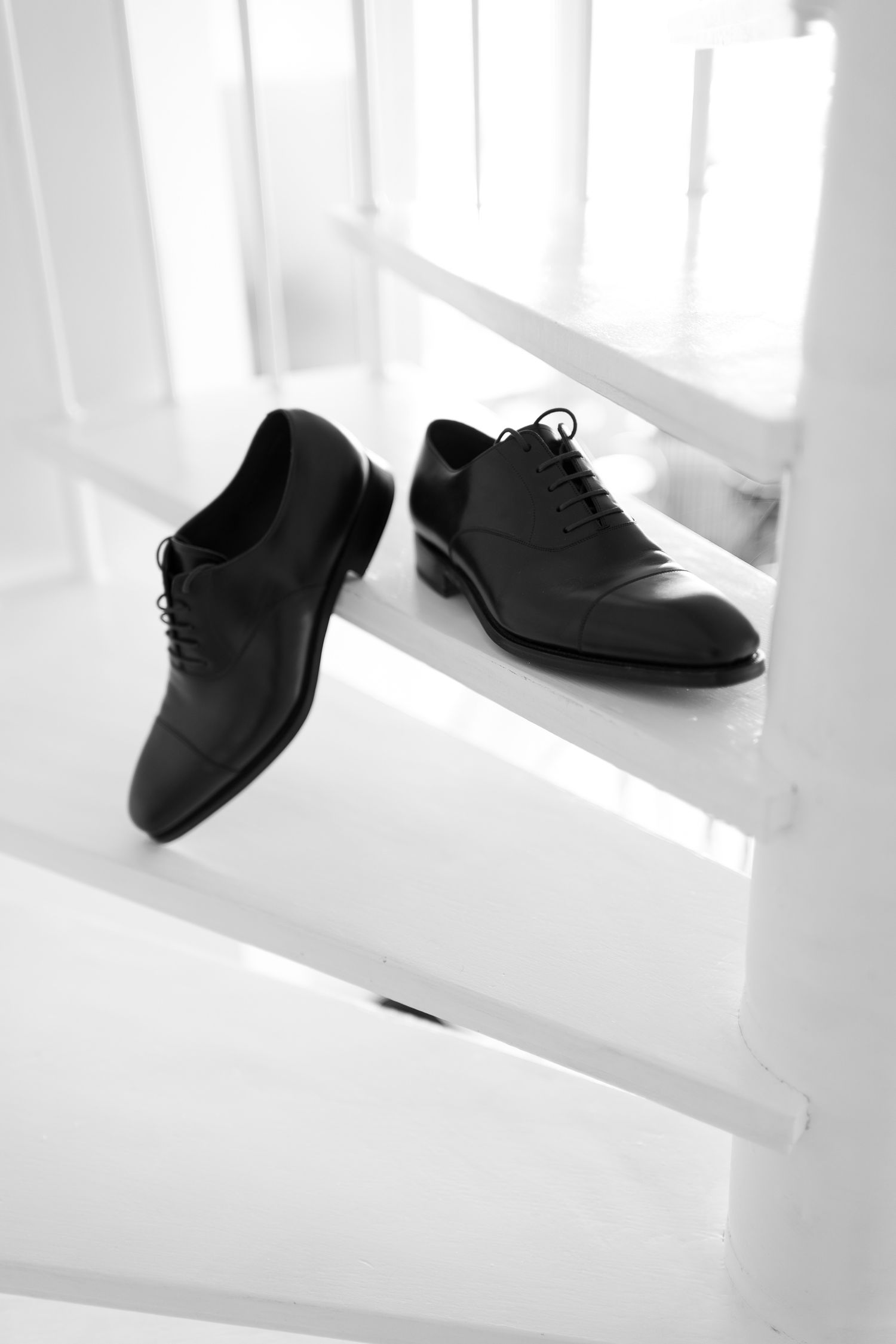 Black leather oxford dress shoes with cap toe and lace-up detail placed on white wooden stairs.