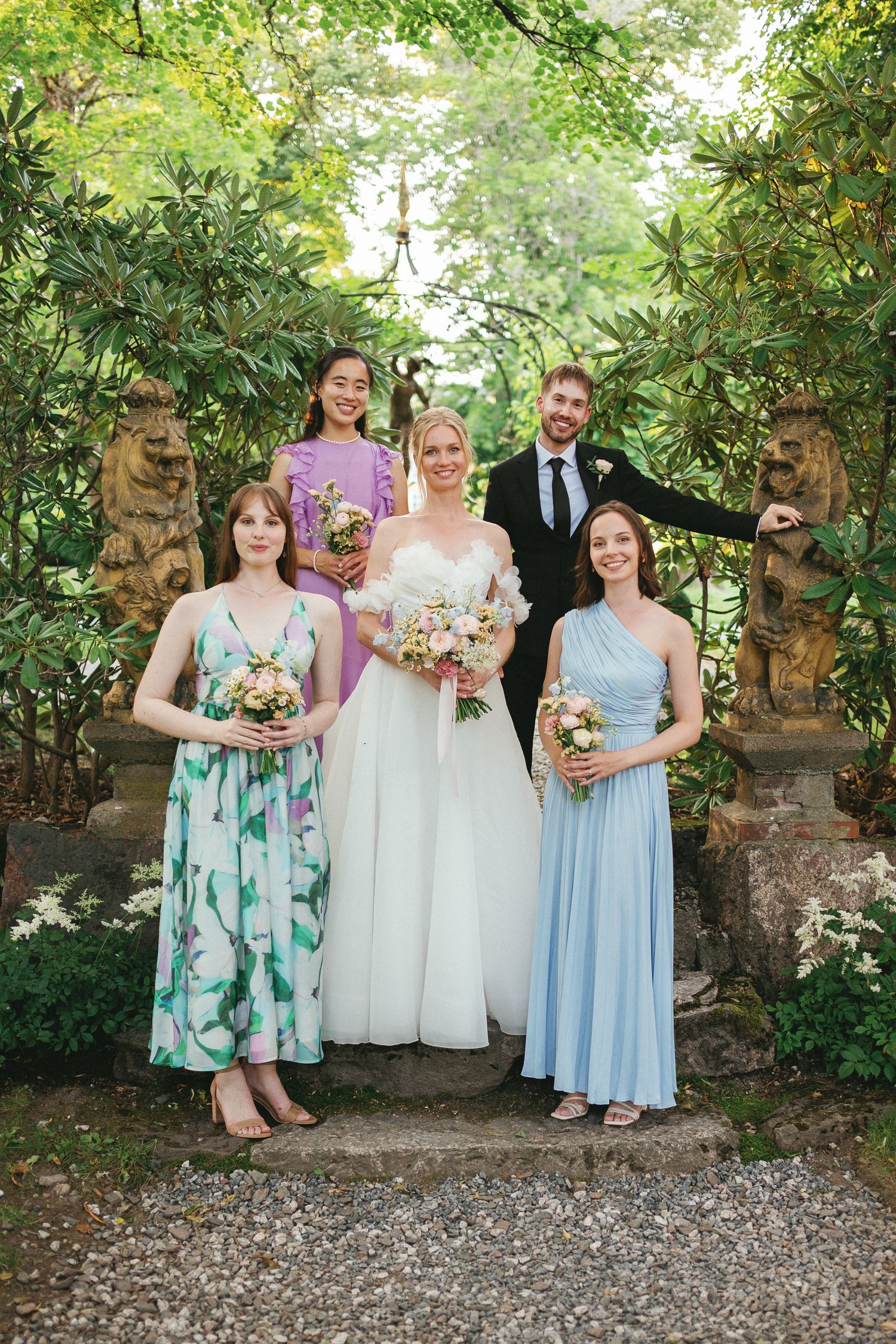 Bride in white gown poses with bridesmaids in colorful dresses and groomsman in lush garden setting.