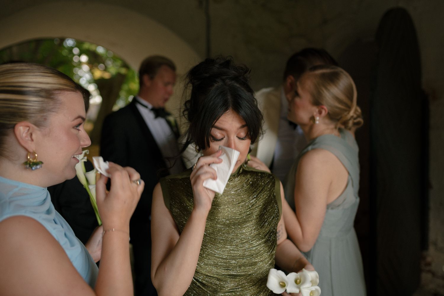 Emotional wedding guest in green dress dabbing tears with tissue, surrounded by bridesmaids in blue dresses at elegant venue.